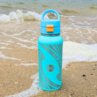 Turquoise water bottle with a handle on a sandy beach with ocean waves.