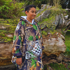 wearing a camo jacket standing in a natural setting with logs and greenery.
