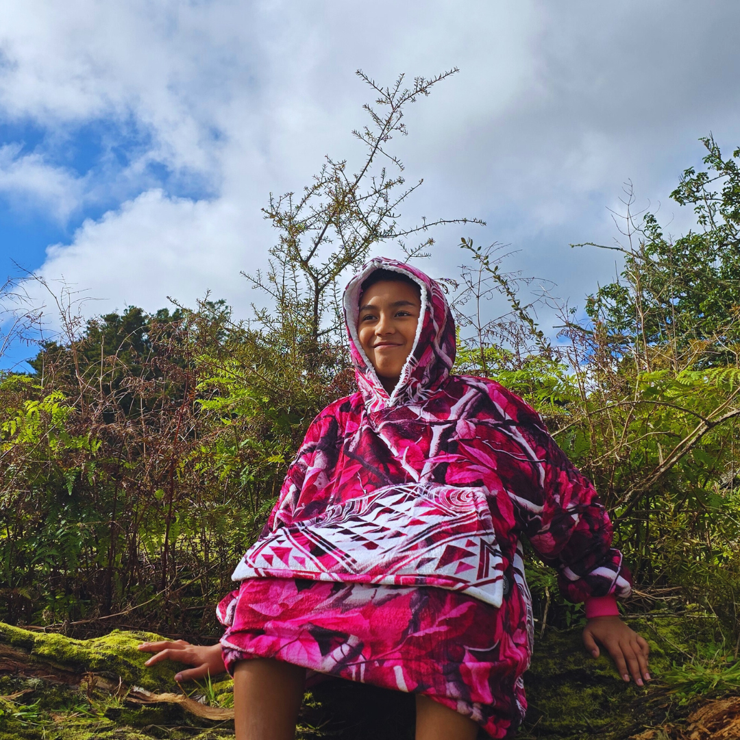Person wearing a pink camo poncho sitting in a forest with trees and blue sky in the background