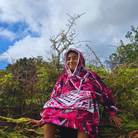 Person wearing a pink camo poncho sitting in a forest with trees and blue sky in the background